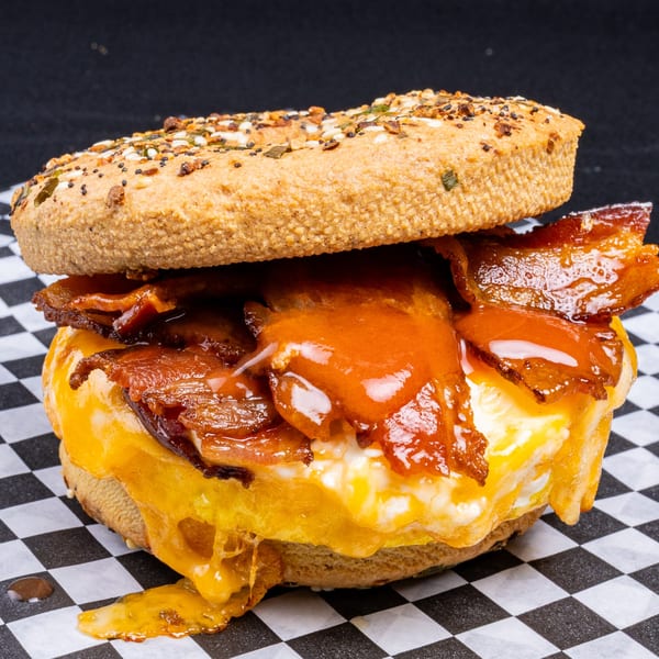 Everything bagel sandwich with crispy bacon, melted cheese, and a fried egg on checkered paper.