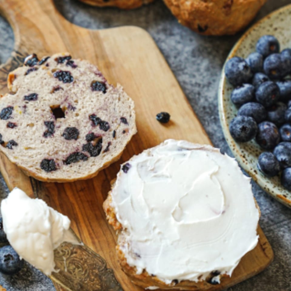 Blueberry bagel, sliced and spread with cream cheese, next to a bowl of fresh blueberries.